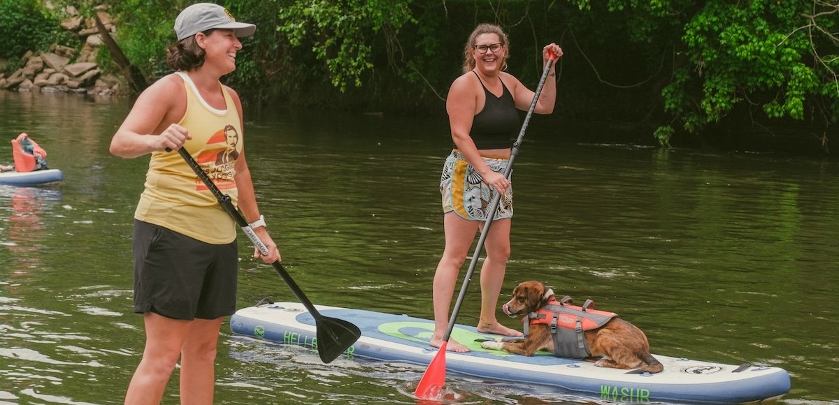 Two women paddle boarding on river. A dog is laying on one of the paddle boards.